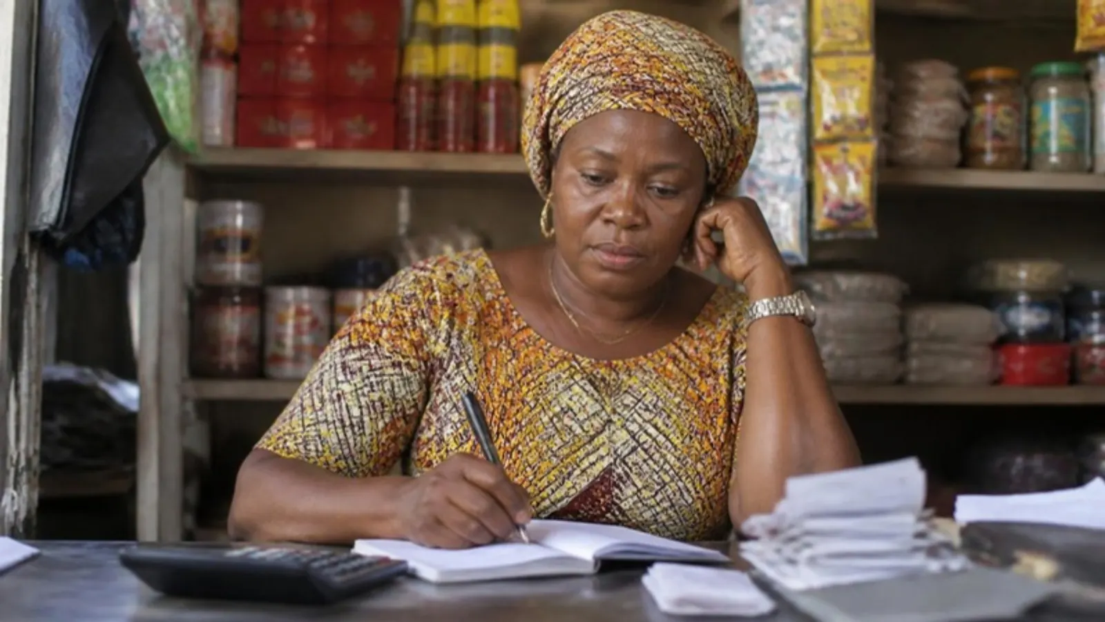 A Nigerian shop owner writing figures in a notebook beside a calculator at her counter